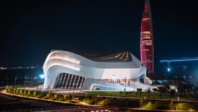 Night view of a futuristic white curved building with layered lighting, against a tall red-lit skyscraper at night.
