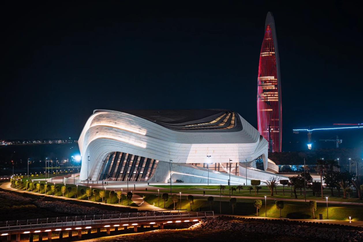Night view of a futuristic white curved building with layered lighting, against a tall red-lit skyscraper at night.