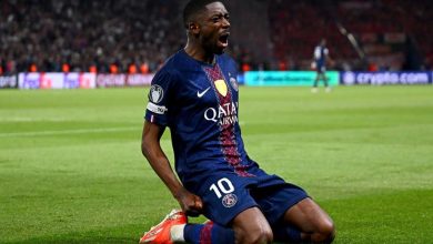 PSG soccer player kneels on the field, shouting in celebration, wearing a navy kit with Qatar Airways sponsor.