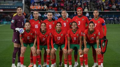 Moroccan women's national football team posing for a group photo on the field, red jerseys with green trim and numbers, goalkeeper in purple on the left.