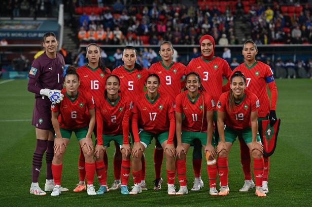 Moroccan women's national football team posing for a group photo on the field, red jerseys with green trim and numbers, goalkeeper in purple on the left.