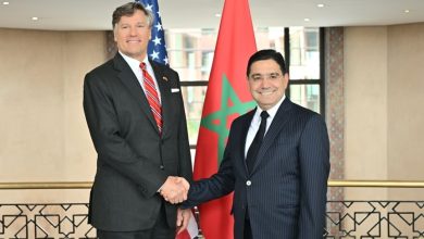 Two men in suits shake hands in front of US and Morocco flags, smiling in a formal setting.