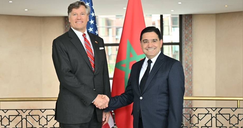 Two men in suits shake hands in front of US and Morocco flags, smiling in a formal setting.