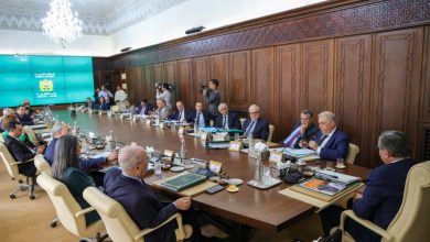 Large conference room with a long wooden table, businesspeople in suits, microphones and documents; teal emblem screen on the left.