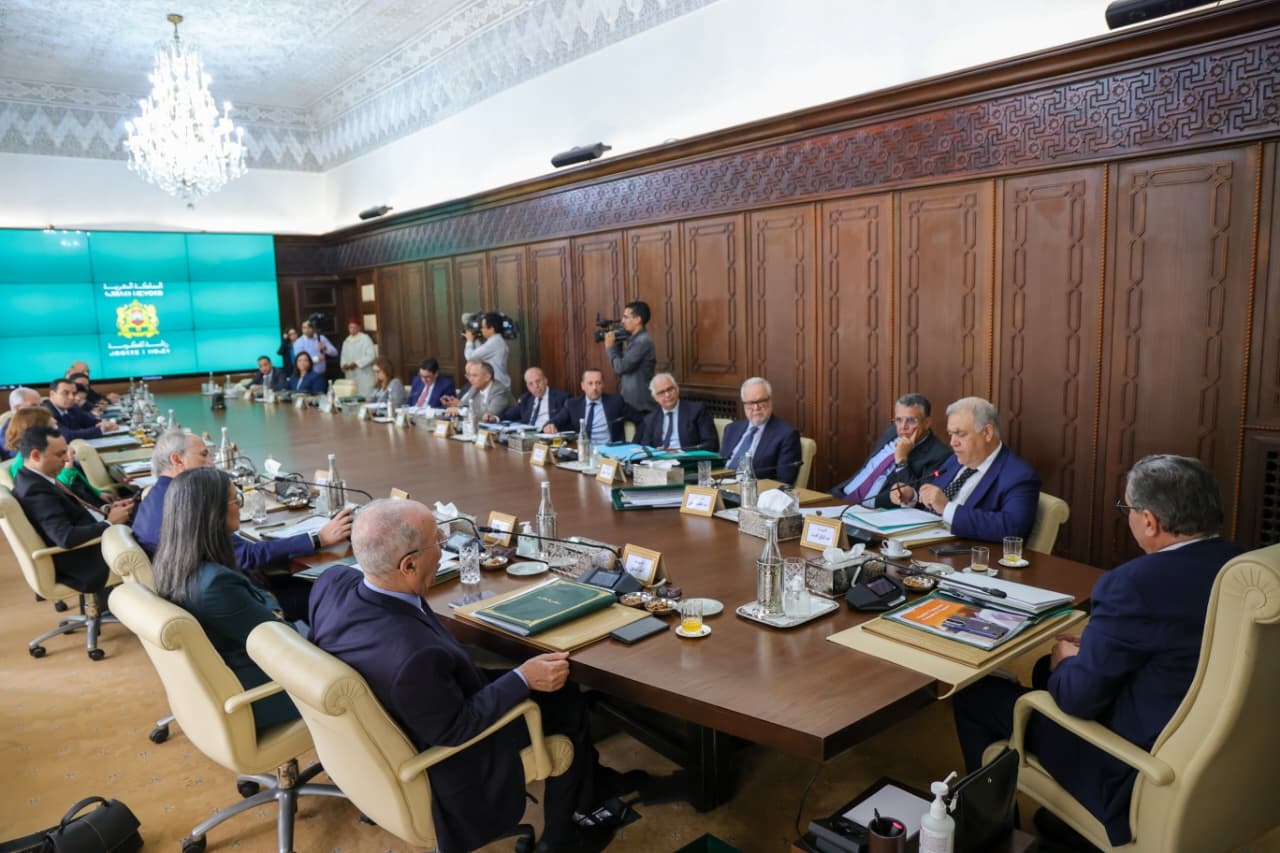 Large conference room with a long wooden table, businesspeople in suits, microphones and documents; teal emblem screen on the left.