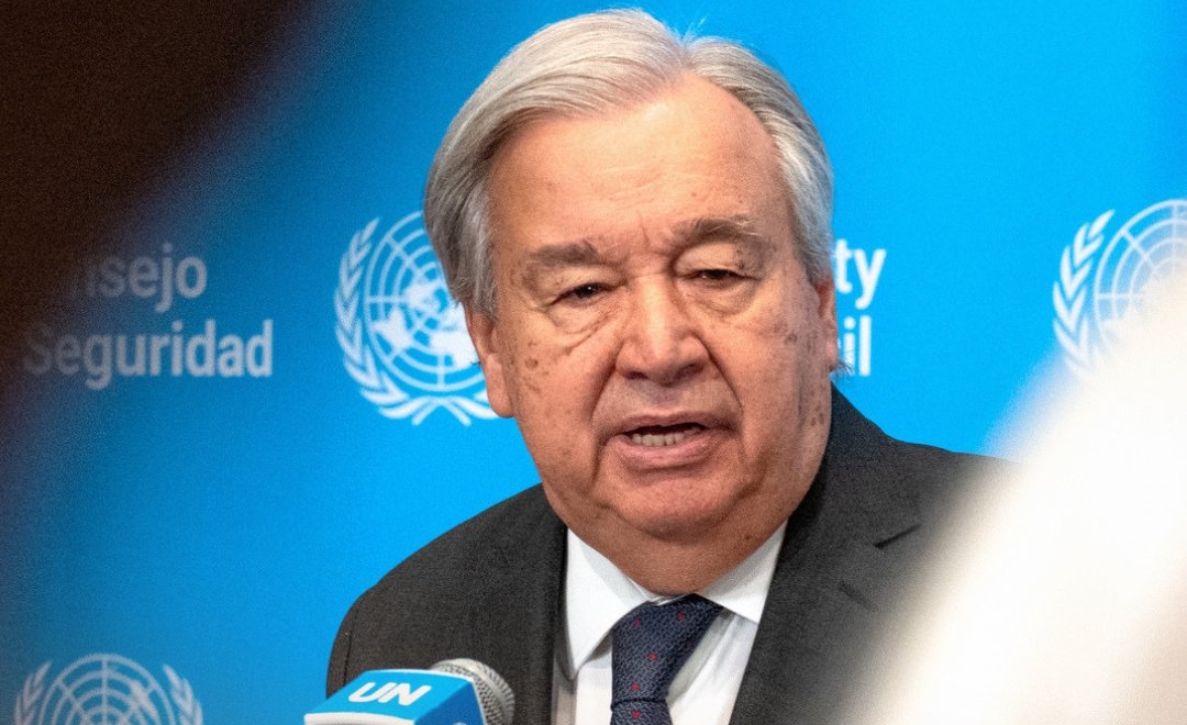 Older man in a suit speaking at a UN press conference, with a blue UN backdrop and microphone labeled 'UN' in the foreground