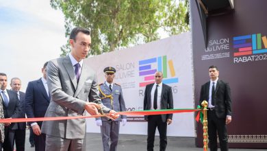 Man in a gray suit cuts a red ribbon at an outdoor ceremony with security personnel and a 'Salon du Livre' backdrop in Rabat.
