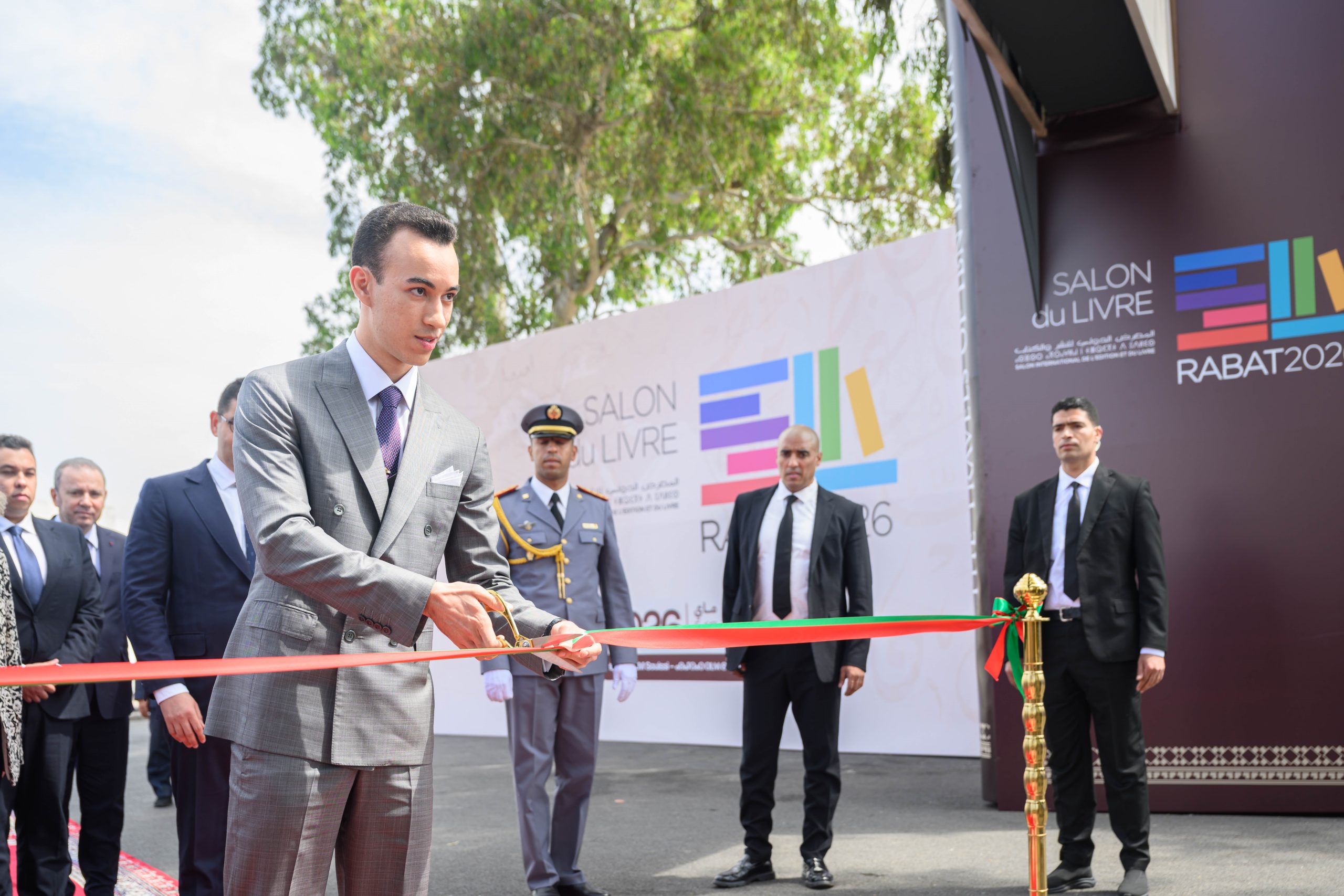 Man in a gray suit cuts a red ribbon at an outdoor ceremony with security personnel and a 'Salon du Livre' backdrop in Rabat.