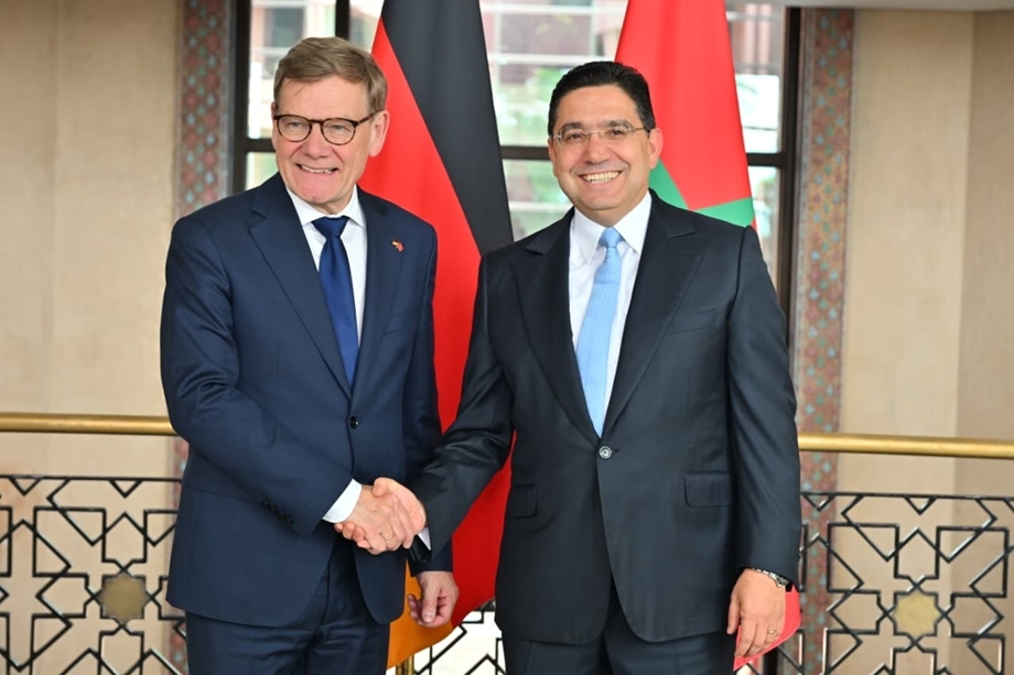Two men in dark suits shake hands in front of red and black flags, standing in a formal indoor setting.