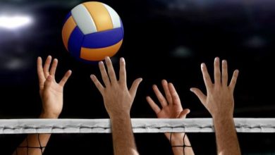 Volleyball players' hands reach over the net to block a yellow, blue, and white ball in a dim arena.