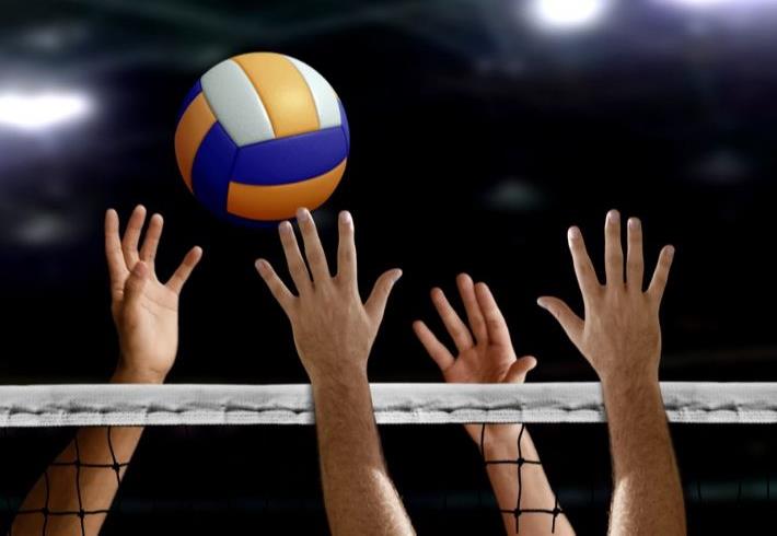 Volleyball players' hands reach over the net to block a yellow, blue, and white ball in a dim arena.