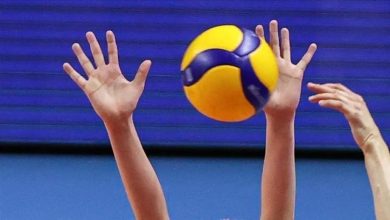 Two hands reaching up to block or receive a yellow-and-blue volleyball mid-air during a game