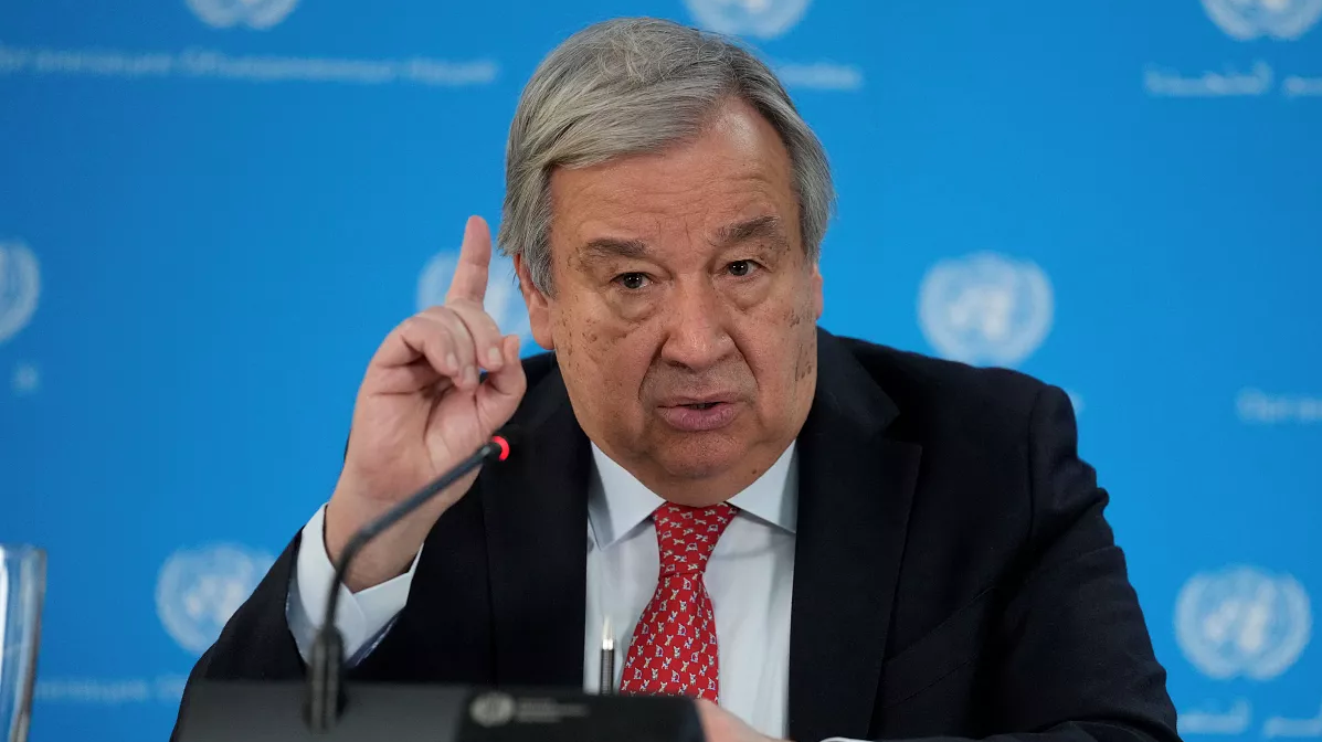 UN official with gray hair in a dark suit and red tie raises his index finger while speaking into a microphone at a briefing, blue UN backdrop behind him.
