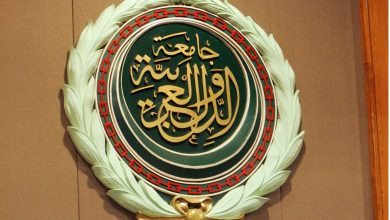 Circular emblem with gold Arabic calligraphy on dark green, bordered by a red chain pattern and pale laurel leaves.