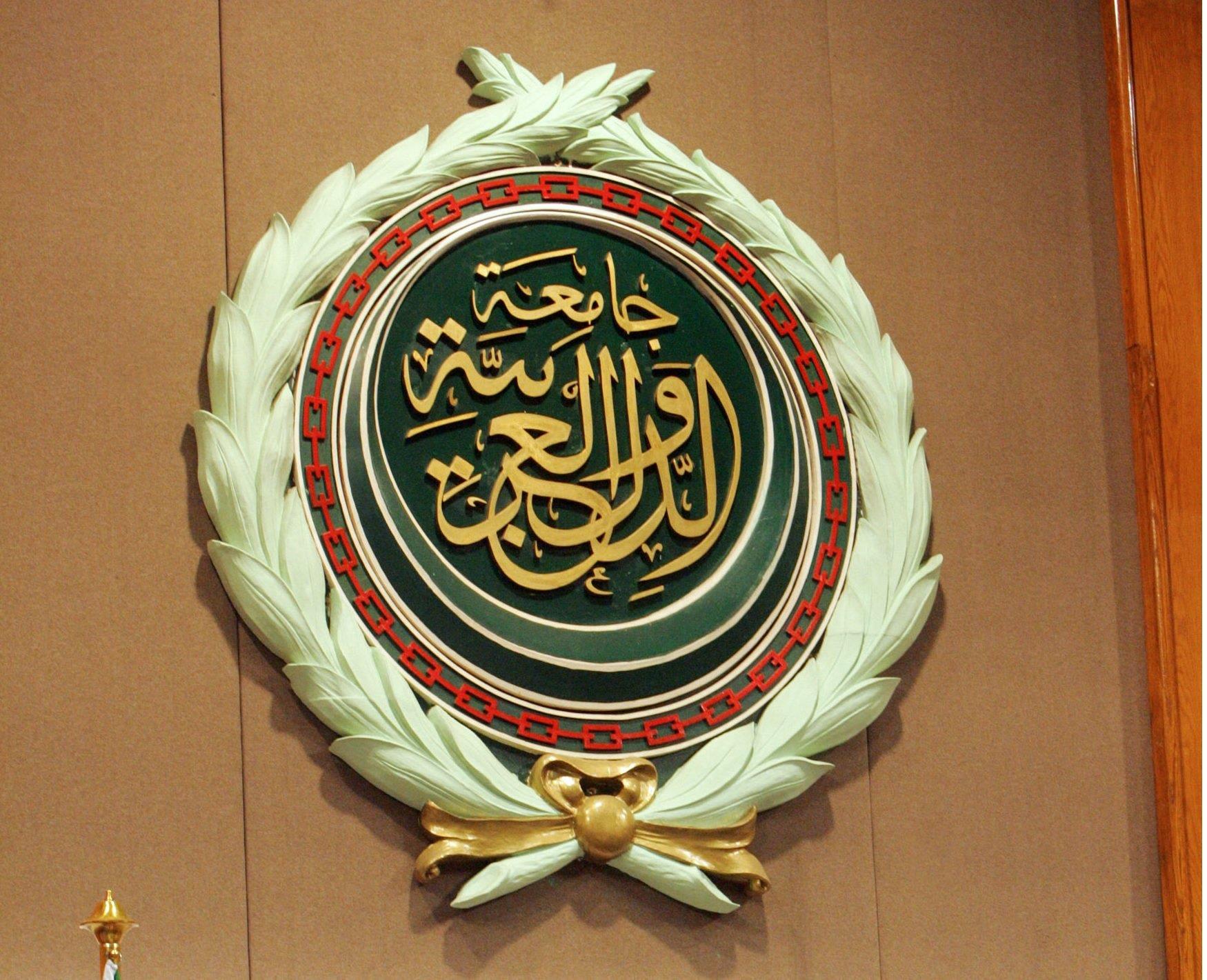Circular emblem with gold Arabic calligraphy on dark green, bordered by a red chain pattern and pale laurel leaves.