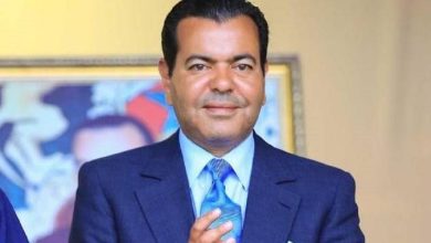 Man in a navy suit with light blue shirt and blue tie, standing indoors with a painting in the background.