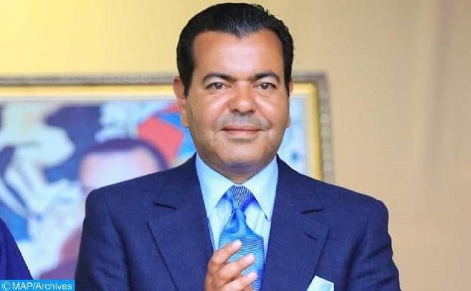 Man in a navy suit with light blue shirt and blue tie, standing indoors with a painting in the background.