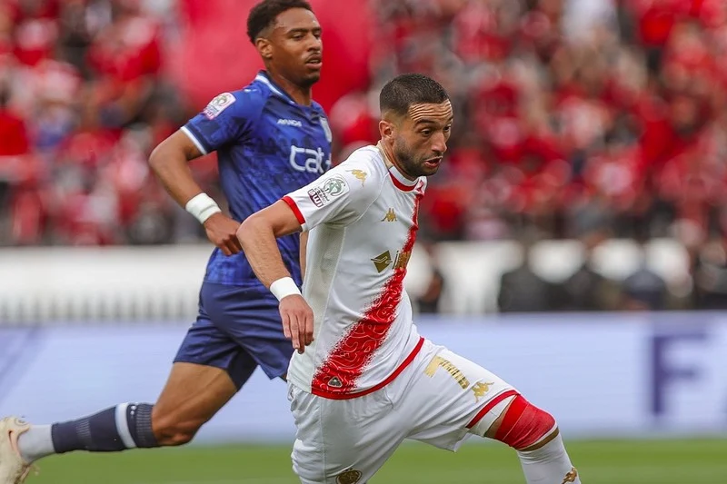Two soccer players chase the ball during a match, one in white with a red diagonal stripe and the other in blue.