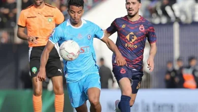 Two soccer players chase the ball during a match: one in light blue kit, the other in dark blue with red pattern, with a referee in orange behind them.