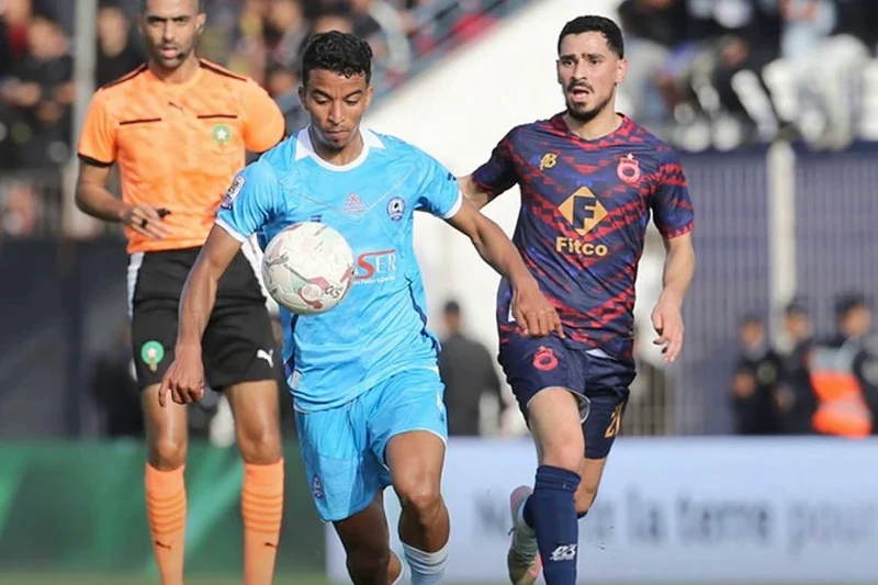 Two soccer players chase the ball during a match: one in light blue kit, the other in dark blue with red pattern, with a referee in orange behind them.