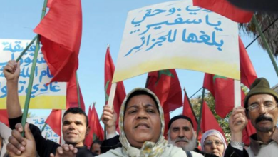 Protesters hold red flags and Arabic banners during a daytime demonstration, faces determined, crowd gathered