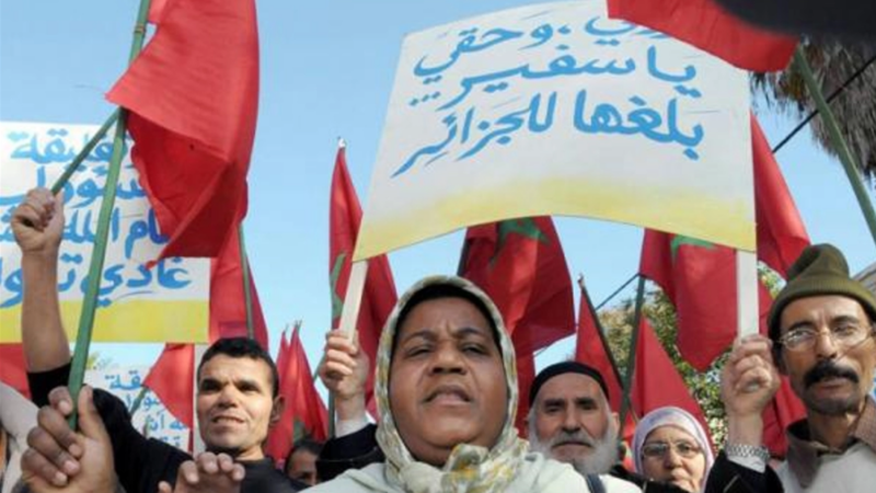 Protesters hold red flags and Arabic banners during a daytime demonstration, faces determined, crowd gathered