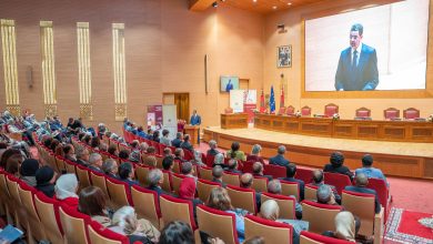 Speaker at a podium addressing a large audience in a formal auditorium with a big screen behind him and flags nearby.