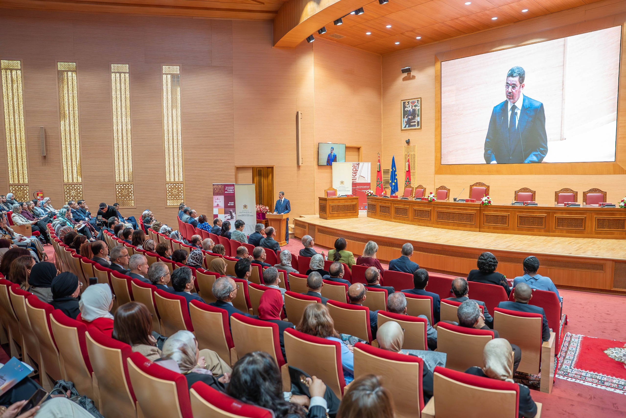 Speaker at a podium addressing a large audience in a formal auditorium with a big screen behind him and flags nearby.