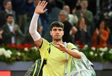 Male tennis player in a yellow shirt waves to the crowd while carrying a gray sports bag off court.