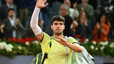 Male tennis player in a yellow shirt waves to the crowd while carrying a gray sports bag off court.