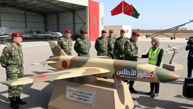 Moroccan military officers in camouflage and red berets gather around a large model missile labeled 'Made in Morocco' as a woman in a safety vest shakes hands with an officer.