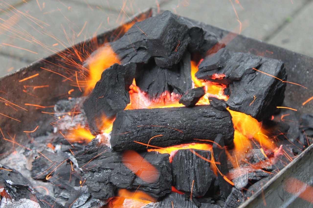 Glowing orange embers and black charcoal briquettes in a metal fire pit with sparks flying around.