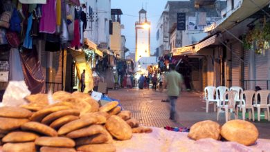 Twilight street market with bread piling in the foreground, shop stalls along both sides, and a lone man walking away among white plastic chairs to the right