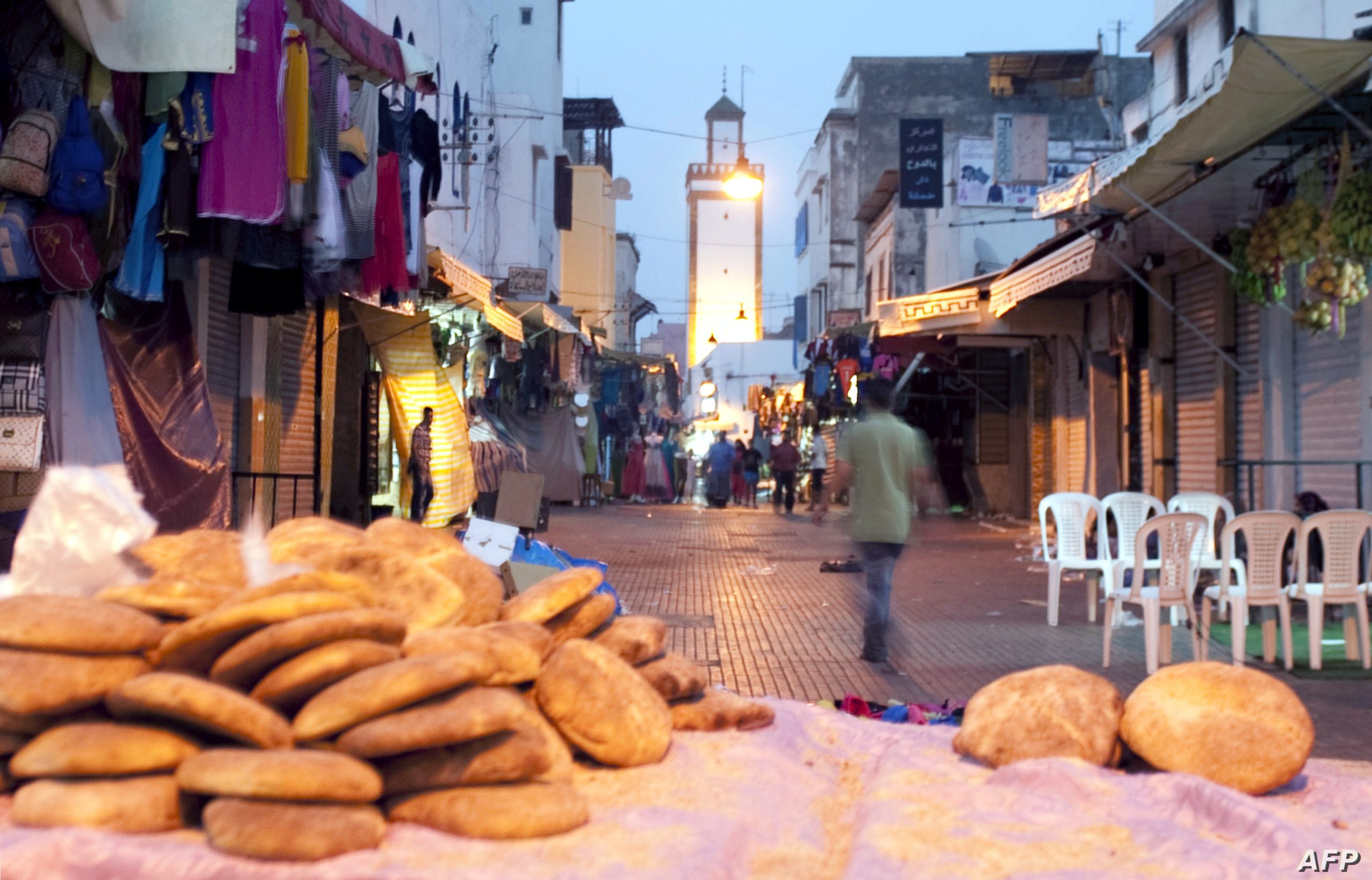 Twilight street market with bread piling in the foreground, shop stalls along both sides, and a lone man walking away among white plastic chairs to the right