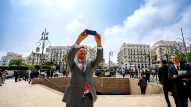 Man in a light gray suit and red tie holds a phone overhead to take a selfie in a busy city square with onlookers and white buildings behind him.