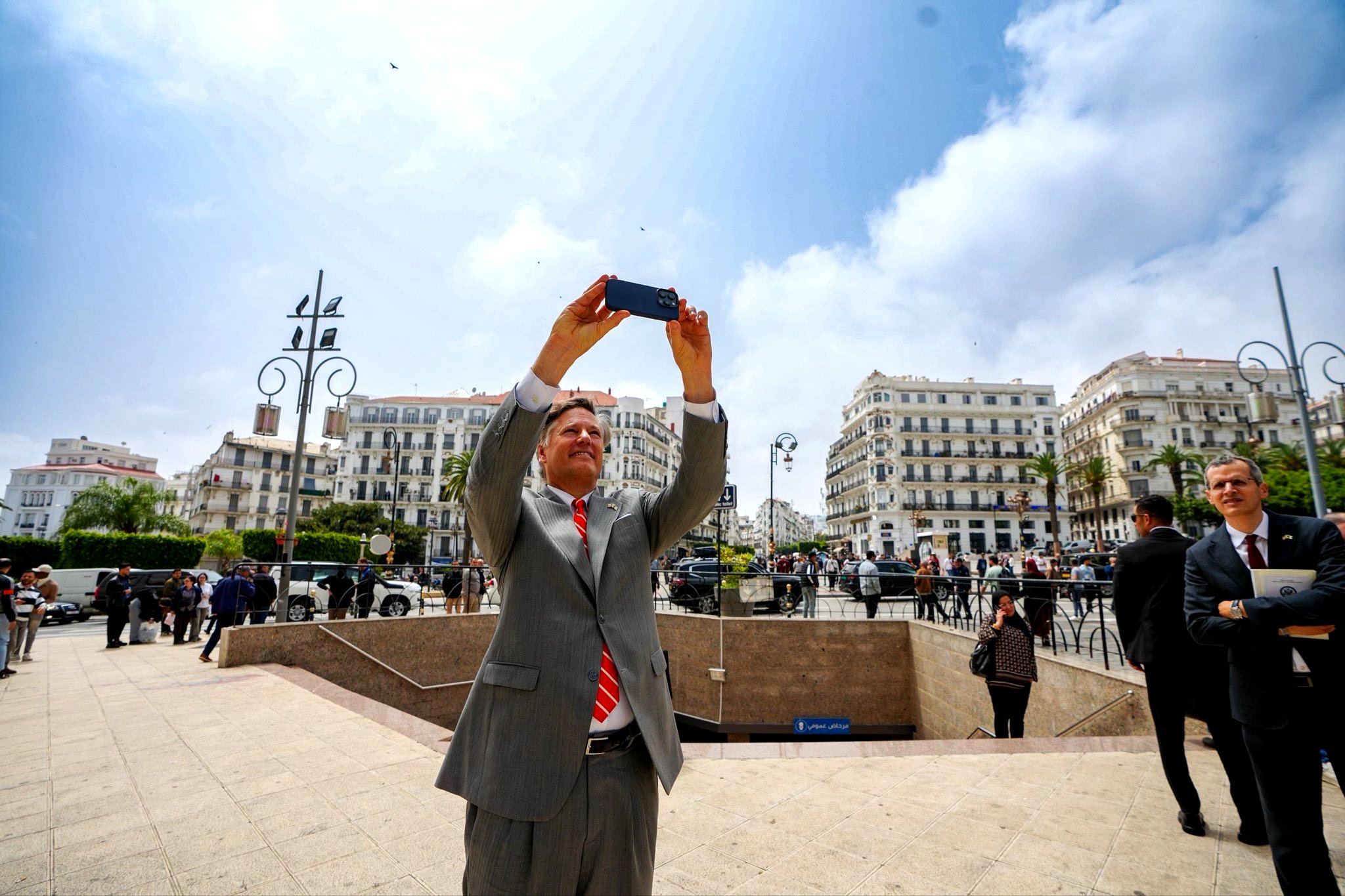 Man in a light gray suit and red tie holds a phone overhead to take a selfie in a busy city square with onlookers and white buildings behind him.
