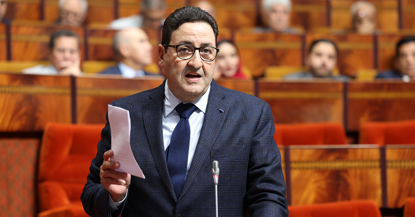 Man in a dark suit and glasses speaks at a lectern in a parliamentary chamber, holding papers while addressing an audience.