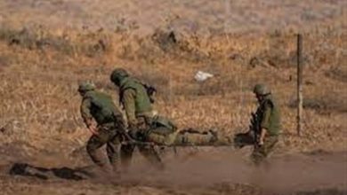 Four soldiers in desert uniforms carry a wounded comrade on a stretcher across a dusty field.