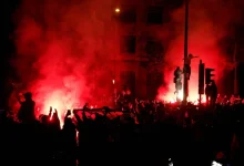 Nighttime protest with a dense crowd and red smoke from flares lighting the street.