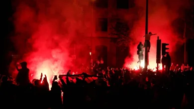 Nighttime protest with a dense crowd and red smoke from flares lighting the street.