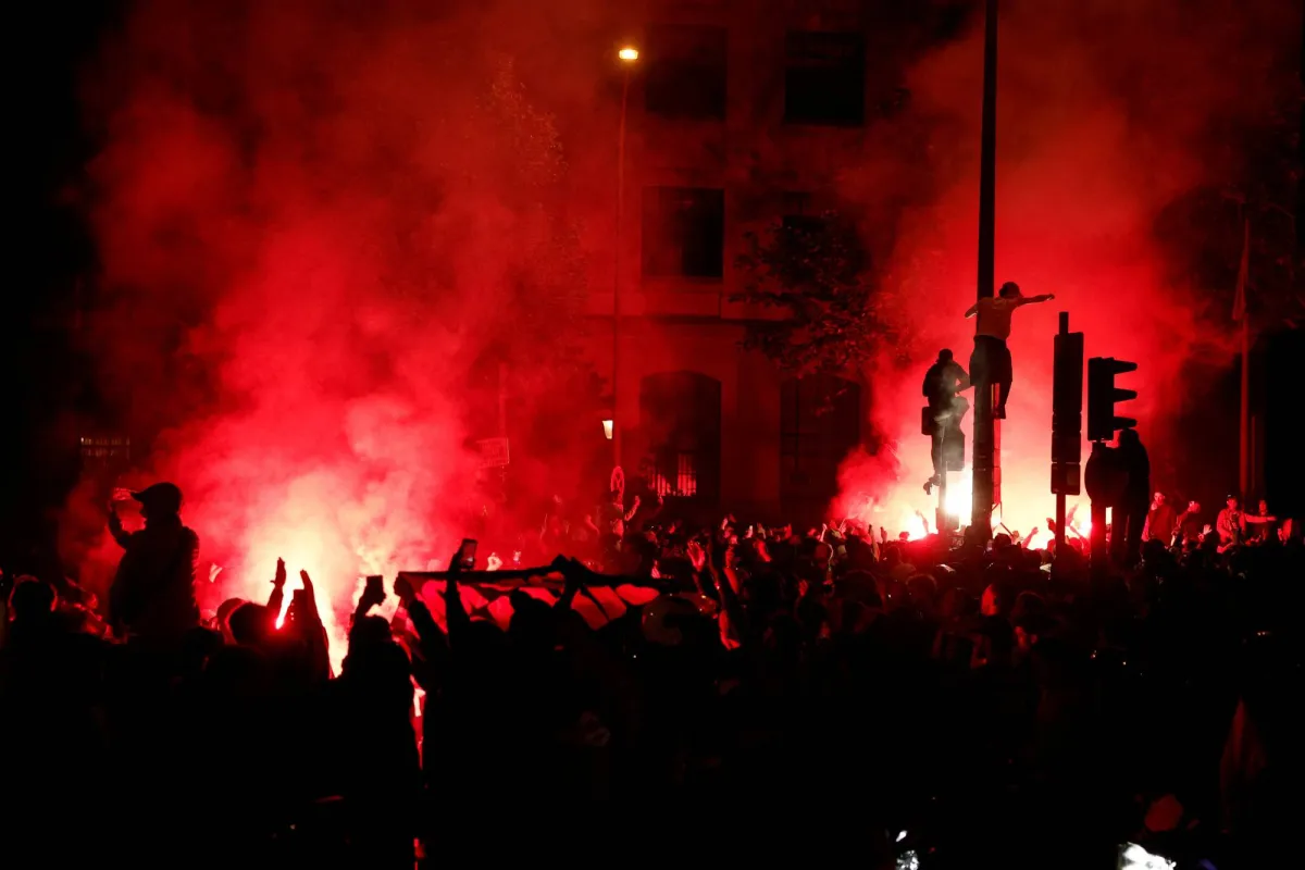 Nighttime protest with a dense crowd and red smoke from flares lighting the street.