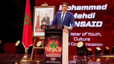 11-1024x683 | MCG24 | موقع إلكتروني مغربي شامل متجدد على مدار الساعة Speaker in a blue suit at a podium on a red-lit stage, with a Moroccan flag and framed portrait beside him to the left.