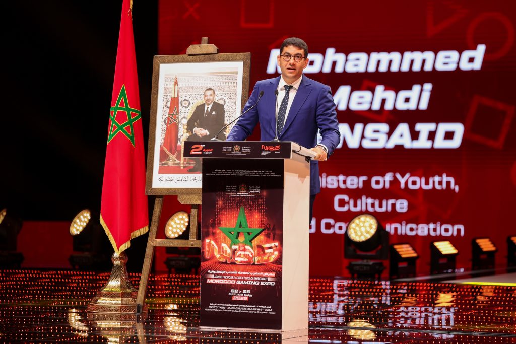 11-1024x683 | MCG24 | موقع إلكتروني مغربي شامل متجدد على مدار الساعة Speaker in a blue suit at a podium on a red-lit stage, with a Moroccan flag and framed portrait beside him to the left.