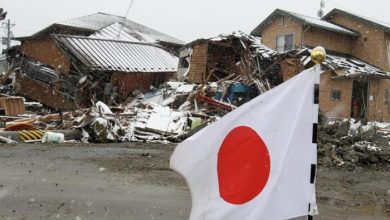 Japanese flag flapping in wind in front of collapsed buildings and debris on a snowy street.