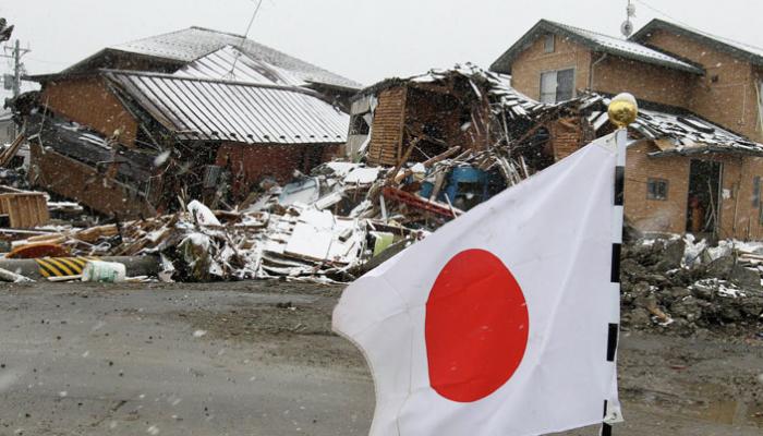 Japanese flag flapping in wind in front of collapsed buildings and debris on a snowy street.