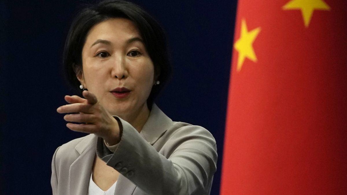 Asian woman in a light gray blazer speaks and gestures, with a Chinese flag in the background.