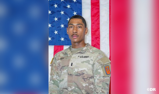 U.S. Army soldier in camouflage uniform standing in front of a large American flag in a posed portrait.