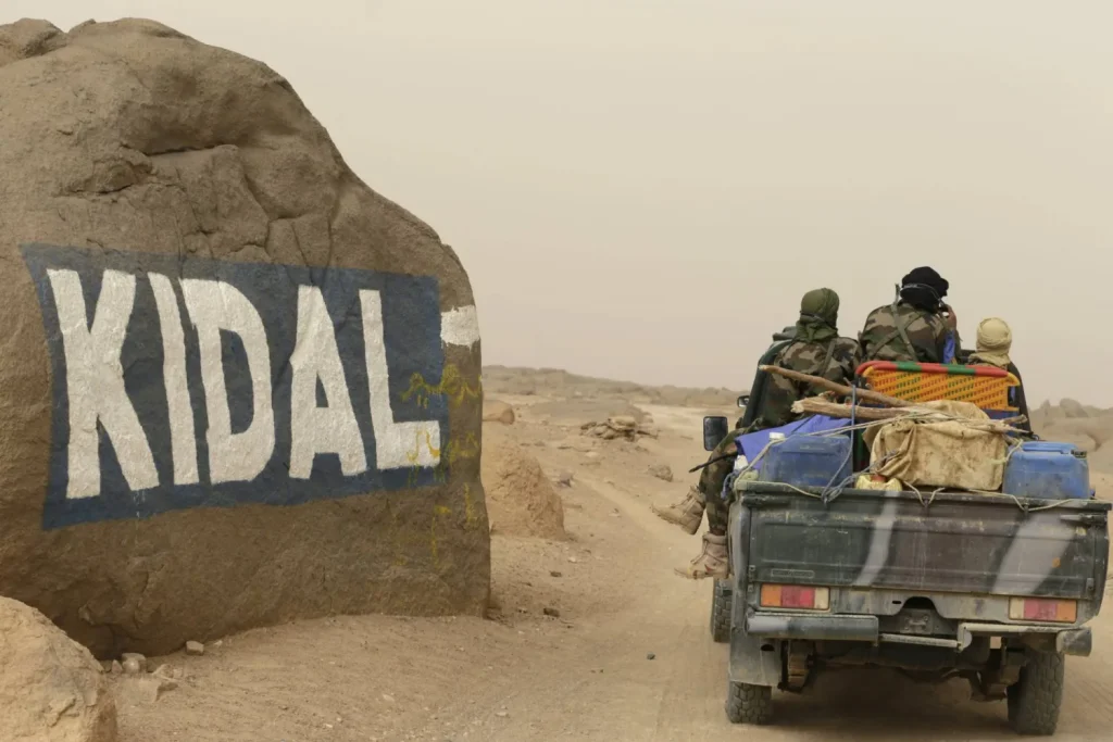 1512121jpeg | MCG24 | موقع إلكتروني مغربي شامل متجدد على مدار الساعة Desert road scene with a pickup truck carrying armed people in camo, driving past a large rock painted with the word 'KIDAL'.