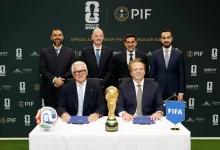 Six men in suits at a signing ceremony with the FIFA World Cup 2026 trophy on the table, ball to the left and a blue FIFA flag to the right.
