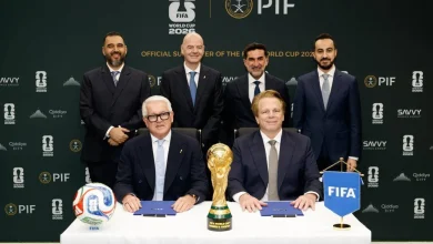 Six men in suits at a signing ceremony with the FIFA World Cup 2026 trophy on the table, ball to the left and a blue FIFA flag to the right.